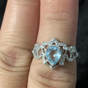 May include: A silver ring featuring a pear-shaped, light blue gemstone at the center. The ring has a decorative, openwork design with small, clear accent stones. The ring is shown on a finger.