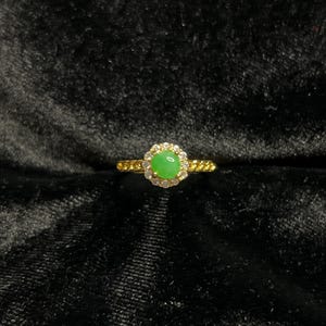 May include: A gold ring featuring a round, light green gemstone surrounded by small, clear stones. The ring's band has a textured, chain-like design. The ring is set against a dark, textured background.