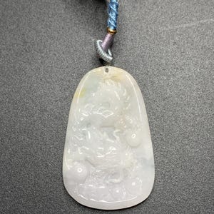 May include: A white jade pendant with a carved dragon design. The pendant is oval-shaped and hangs from a cord with blue, purple, and gold accents. The dragon is intricately detailed, with a smooth, polished finish.