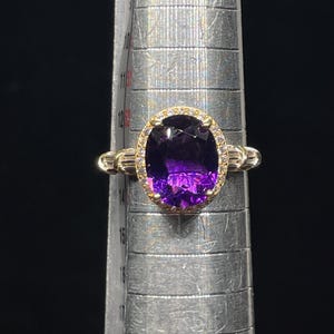 May include: A gold ring with a large, oval-cut purple gemstone, surrounded by smaller clear stones. The ring is set on a silver measuring tool, with a dark background. The ring's band has a textured design.