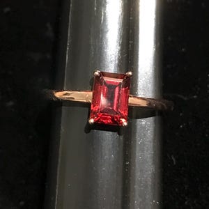 May include: A gold ring featuring a rectangular emerald-cut red gemstone set in a four-prong setting. The band is thin and polished, showcasing a vibrant, deep red stone that sits prominently at the center of the metal ring structure.