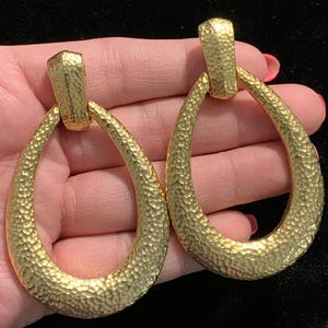 May include: A pair of gold-colored, textured teardrop earrings. The earrings have a hammered metal appearance and a rectangular top piece. The teardrop shape is elongated and has a slightly irregular texture, giving them a vintage look.