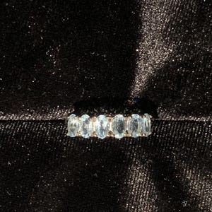 May include: A ring featuring a row of seven oval-cut, light blue gemstones set in a metallic band. The stones are arranged horizontally, displaying a pale, translucent blue hue that contrasts with the dark, textured fabric background.