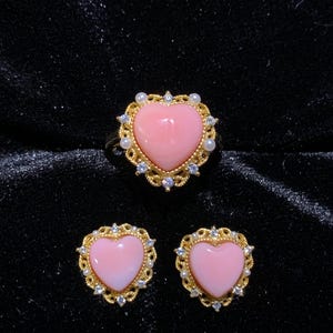 May include: A gold-toned jewelry set consisting of a ring and two stud earrings. Each piece features a smooth, light pink heart-shaped center stone surrounded by a decorative gold filigree frame accented with small white pearls and clear crystals.
