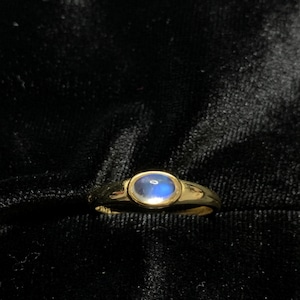 May include: A gold ring with a blue oval-shaped stone. The ring is set against a black background, highlighting the polished gold band and the iridescent quality of the gemstone. The ring's design is simple and elegant.