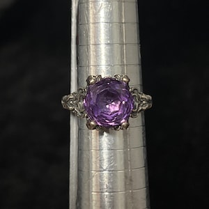 May include: A silver ring with a large, faceted, round purple gemstone. The ring has an ornate, filigree design around the gemstone and on the band. The ring is displayed on a silver ring sizer.