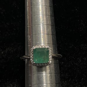 May include: A silver ring with a square, emerald-cut green gemstone, surrounded by a halo of small, clear stones. The ring is on a silver ring sizer, set against a dark background.