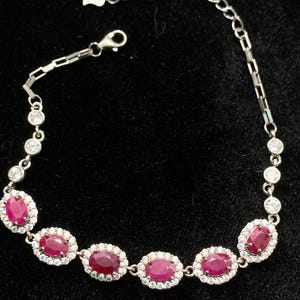 May include: A silver bracelet featuring oval-cut, deep red gemstones surrounded by small, clear stones. The bracelet has a delicate chain and a secure clasp. The gemstones are evenly spaced along the bracelet's length, creating a classic and elegant design.