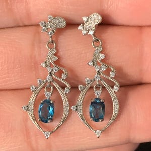 May include: A pair of silver-toned drop earrings. Each earring features a blue gemstone within an oval frame, accented with small clear stones. The design includes a decorative top and a flowing, openwork pattern.