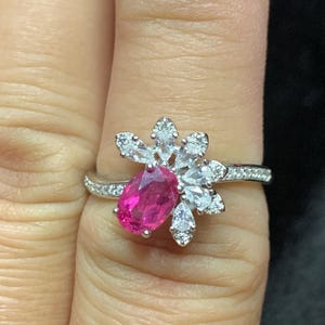 May include: A silver ring with a pink oval gemstone surrounded by smaller clear gemstones in a floral design.