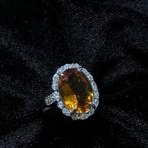 May include: A silver ring with a large oval-shaped orange gemstone surrounded by a halo of smaller clear gemstones.
