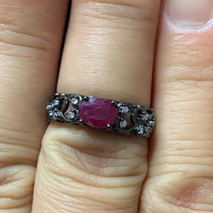 May include: A dark metal ring featuring an oval-cut ruby gemstone. The ring is adorned with small, clear gemstones and an ornate filigree design. The ruby is a vibrant red color, and the ring is set against a neutral background.