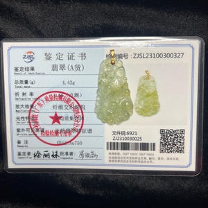 May include: A jade pendant with a gold bail is displayed next to a smaller jade pendant. The pendants are light green and are on a certificate with Chinese text. The certificate has a red stamp and text in Chinese and English.