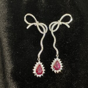 May include: A pair of silver earrings with a bow design and a teardrop-shaped ruby gemstone surrounded by smaller diamonds.