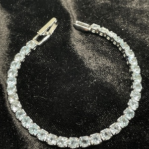 May include: A silver bracelet with a row of square, light blue gemstones. The bracelet is unclasped, showing the clasp mechanism. The background is a dark, textured fabric.