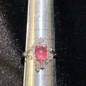 May include: A silver-toned ring featuring a central pear-shaped pink gemstone surrounded by a cluster of small, clear, rectangular and round-cut stones. The ring is positioned on a metal ring mandrel marked with imperial measurement lines.