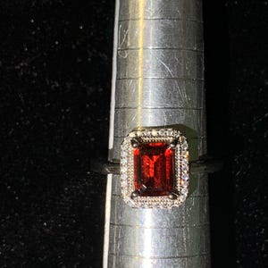 May include: A ring featuring a rectangular, deep red gemstone set in a silver-toned metal band. The center stone is surrounded by a halo of small, clear, sparkling stones. The ring is positioned on a silver metal ring mandrel with visible size markings.