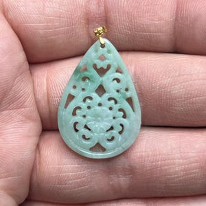 May include: A jade pendant with a teardrop shape and intricate openwork design. The pendant features a floral motif in the center and is suspended from a gold-colored bail. The jade has a pale green hue.