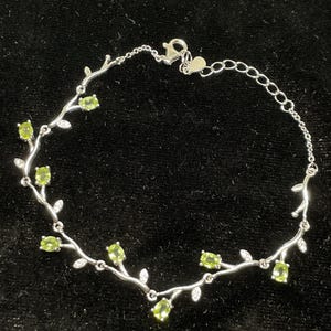 May include: A silver bracelet featuring a delicate vine design. The bracelet is adorned with oval-shaped, light green gemstones and small, clear accent stones. The clasp is a lobster claw style. The bracelet is displayed on a black surface.