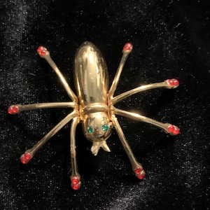 May include: A gold-toned insect brooch with eight slender legs. The insect's body is oval-shaped, with green gemstone eyes and red gemstone accents on the tips of its legs. The brooch is set against a black background.