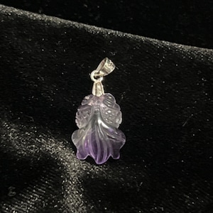 May include: A carved amethyst pendant with a silver bail. The pendant is shaped like a stylized leaf or flower, with a gradient of purple hues. The silver bail is attached to the top of the pendant.