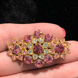 May include: A vintage gold-toned brooch with a cluster of pink and clear rhinestones. The brooch features a rectangular shape with a floral design, showcasing various sizes of round stones. The pink stones vary in shade, creating a gradient effect.