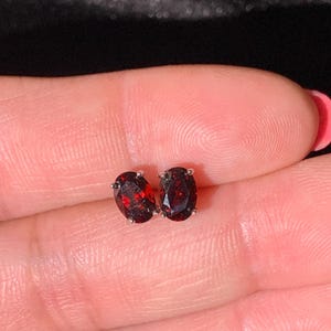 May include: A pair of oval-shaped stud earrings featuring deep red faceted gemstones set in silver-toned metal prongs. The stones exhibit a dark crimson hue with internal reflections, and each is secured by four small metal prongs holding the gem.
