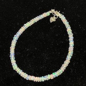 May include: A delicate white opal beaded bracelet with a silver heart charm and clasp. The bracelet is made of small, round opal beads that are strung together on a silver chain.