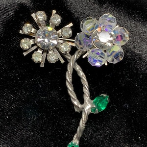 May include: A silver brooch with two crystal flower designs and green leaves. The larger flower is made of faceted crystals and the smaller flower is made of smaller, round crystals.