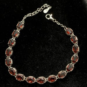 May include: A silver bracelet with oval-shaped, deep red gemstones. The gemstones are set in a silver metal frame and linked together with a delicate chain. The bracelet is displayed against a black background.
