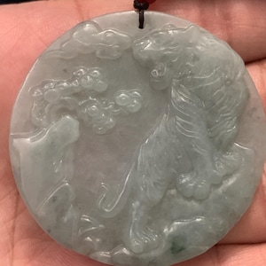 May include: A circular pale green jade pendant featuring a carved tiger standing on a rocky surface with stylized clouds and a tree in the background. The pendant is suspended from a dark brown cord with a small red bead at the top attachment point.
