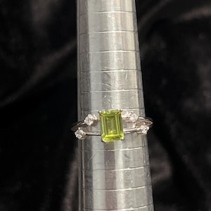 May include: A silver ring with a rectangular, light green gemstone at the center. The ring is flanked by smaller, round, clear gemstones. The ring is displayed on a silver ring sizer against a dark background.