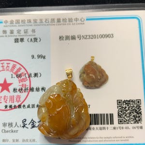 May include: Two jade pendants with gold bails are displayed on a white card. The larger pendant is a mix of yellow and white, with a carved design. The smaller pendant is a similar shape, with a brown hue. The card has Chinese text and a QR code.
