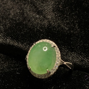 May include: A silver ring featuring a large, oval-shaped, light green gemstone. The gemstone is surrounded by a halo of smaller, clear stones. The ring's band has a delicate, textured design.