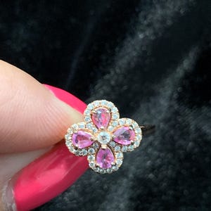 May include: A rose gold ring featuring a four-leaf clover design. Each leaf is a pink pear-shaped gemstone, surrounded by a halo of small, clear stones. A single clear stone sits in the center.