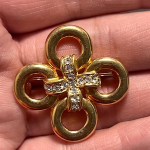 May include: A gold-toned brooch with a four-loop design, each loop connected at the center. The center features a cross-shaped embellishment encrusted with clear rhinestones. The brooch is held in a hand.