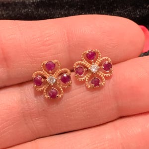 May include: A pair of gold-colored flower-shaped stud earrings. Each earring features four ruby-colored gemstones and a central clear gemstone. The earrings are set against a blurred background.