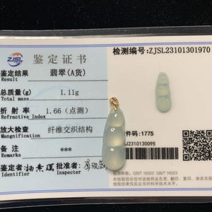 May include: A jade pendant with a gold bail, featuring a three-section design. The pendant is light green and sits on a certificate with Chinese text. The certificate also includes the total mass of the pendant, which is 1.11 grams.
