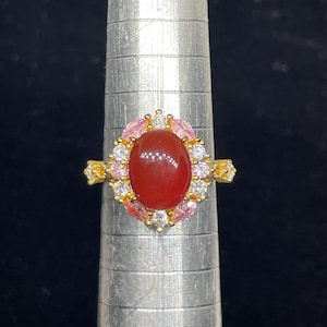 May include: A gold ring with a red oval gemstone surrounded by clear and pink cubic zirconia stones.