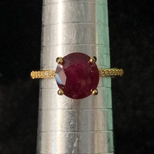 May include: A gold ring featuring a large, round, deep red gemstone held by four prongs. The band is thin, yellow gold, and accented with small, clear stones along the sides. The ring is positioned on a silver metal ring mandrel for sizing.