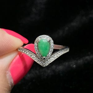 May include: A silver ring featuring a pear-shaped, vibrant green gemstone, surrounded by a halo of small, clear stones. The ring's band has a V-shaped design, also adorned with clear stones, creating a sparkling and elegant appearance.
