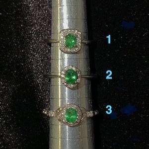 May include: Three silver-toned rings arranged vertically on a metal ring mandrel. Each ring features a central oval-cut green gemstone surrounded by a halo of small clear stones. The rings are labeled with the blue numbers 1, 2, and 3.