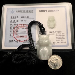 May include: A pale green carved jade cat pendant on a black braided cord, accompanied by a certification card stating a total weight of 14.48g and a US dime coin for scale. The pendant features a seated cat figure with pointed ears and a smooth finish.