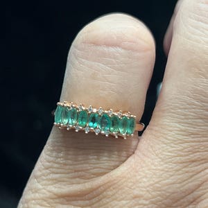May include: Rose gold ring featuring a row of oval-cut emeralds and small, clear accent stones. The emeralds are a vibrant green, and the ring is set against a dark background.