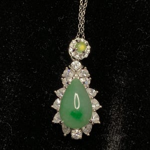 May include: A silver necklace with a pendant featuring a large, pear-shaped green gemstone surrounded by clear, sparkling stones. A smaller, round green stone is set above the pendant. The necklace hangs on a delicate silver chain.