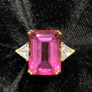 May include: A pink emerald-cut gemstone ring with two smaller triangular gemstones on either side. The ring is set in gold.