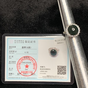 May include: A silver ring with a dark green gemstone center, surrounded by a halo of smaller clear stones. The ring is displayed on a metal ring sizer next to a certificate with Chinese text and a QR code.
