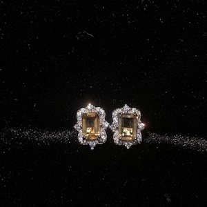 May include: A pair of stud earrings featuring rectangular, emerald-cut yellow gemstones at the center. Each stone is surrounded by a halo of small, clear, round-cut white stones set in a metallic frame, creating a floral-like border design.