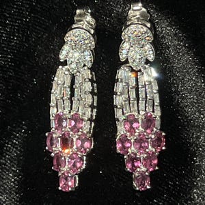 May include: A pair of silver-toned drop earrings. The earrings feature a cluster of pink gemstones at the bottom, with a section of rectangular clear stones above. The top of the earrings have a floral design with clear stones.