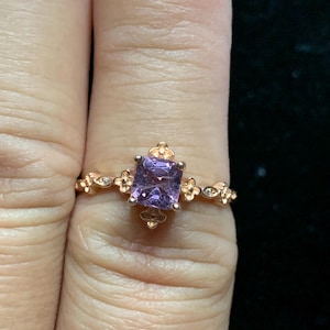 May include: A rose gold ring with a square purple gemstone surrounded by small floral details. The ring is on a finger.